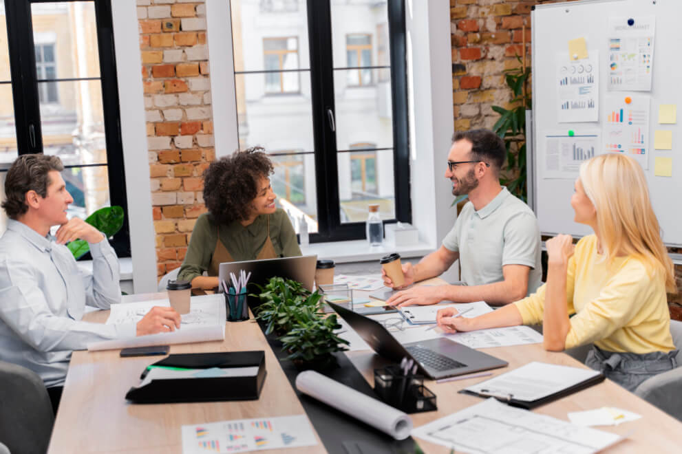 Four people sitting around a wooden desk in a cheery office filled with papers and charts outlining marketing campaign performance.